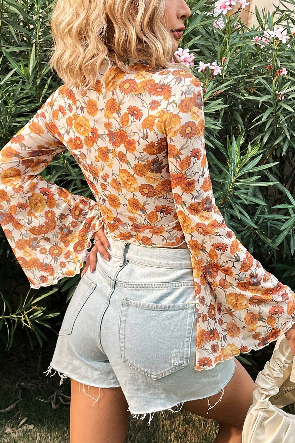 Person wearing a floral blouse and denim shorts standing among green foliage.