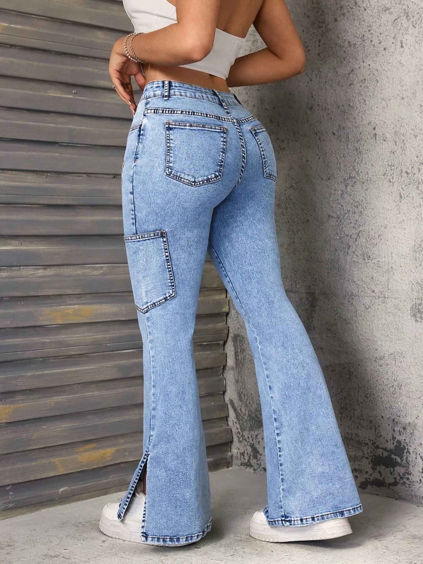 Lady wearing slit star flare leg blue denim jeans paired with a white tank top and white tennis shoes, with walls visible in the background.