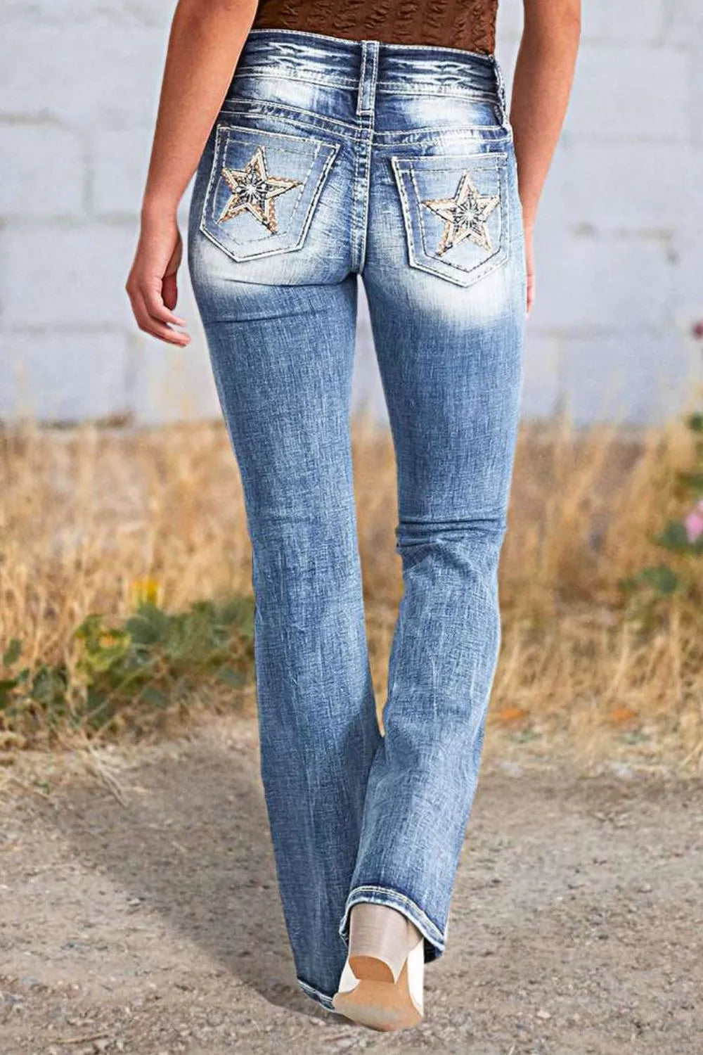 Woman wearing long boot cut acid wash blue jeans with star embroidered pockets, styled with sandals, with dried grass visible in the background.