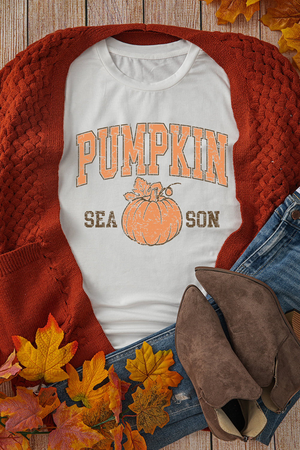 White t-shirt with 'Pumpkin Season' design on a wooden surface with autumn leaves and a red sweater.