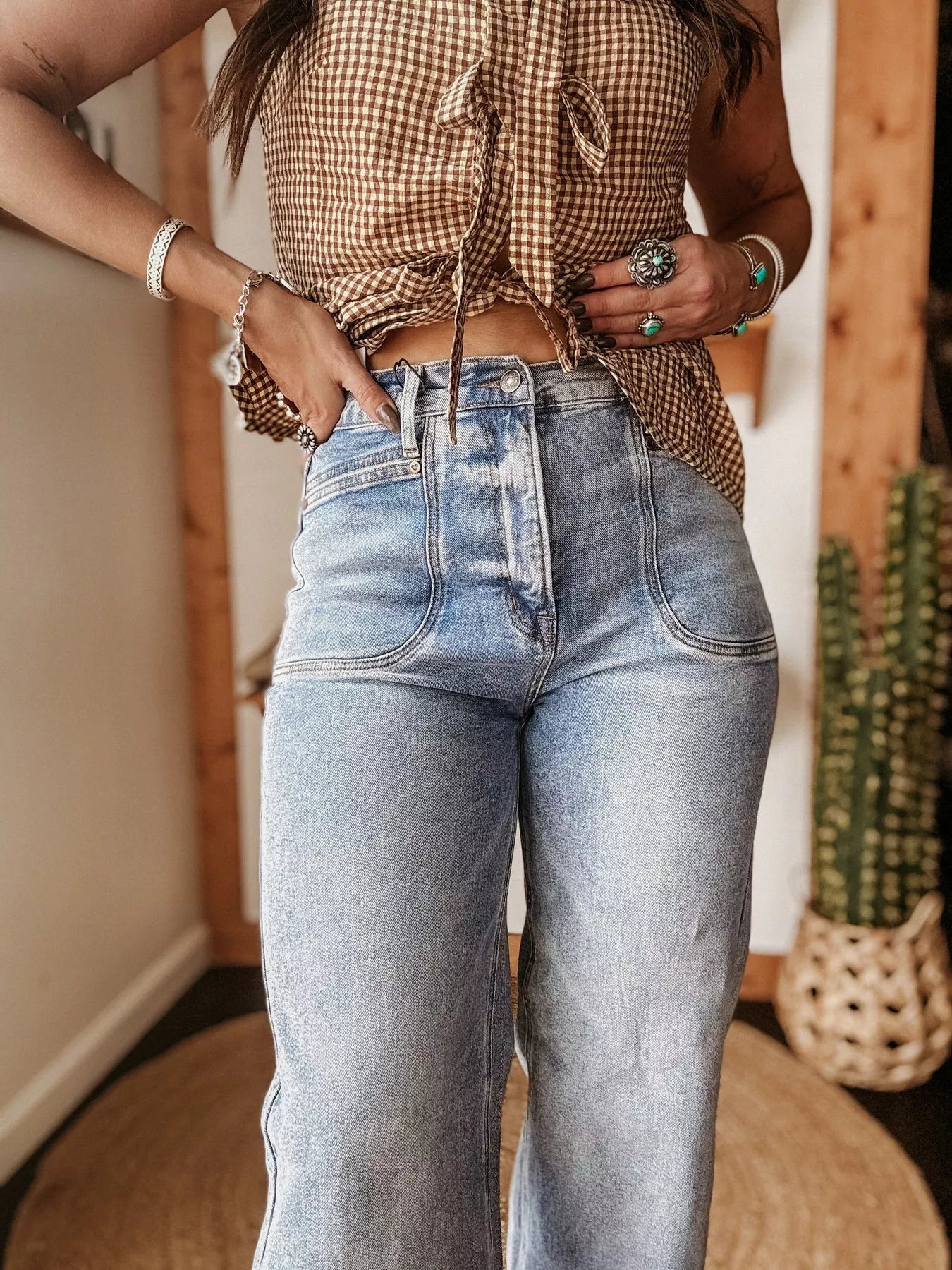 Woman wearing blue denim jeans and a sleeveless bow tie blouse, standing with shoes visible in the background.