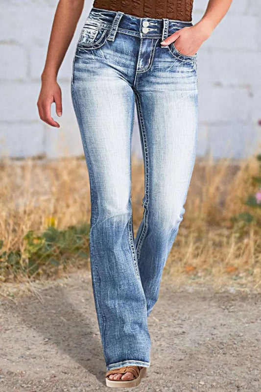 Woman wearing long boot cut acid wash blue jeans with star embroidered pockets, styled with sandals, with dried grass visible in the background.