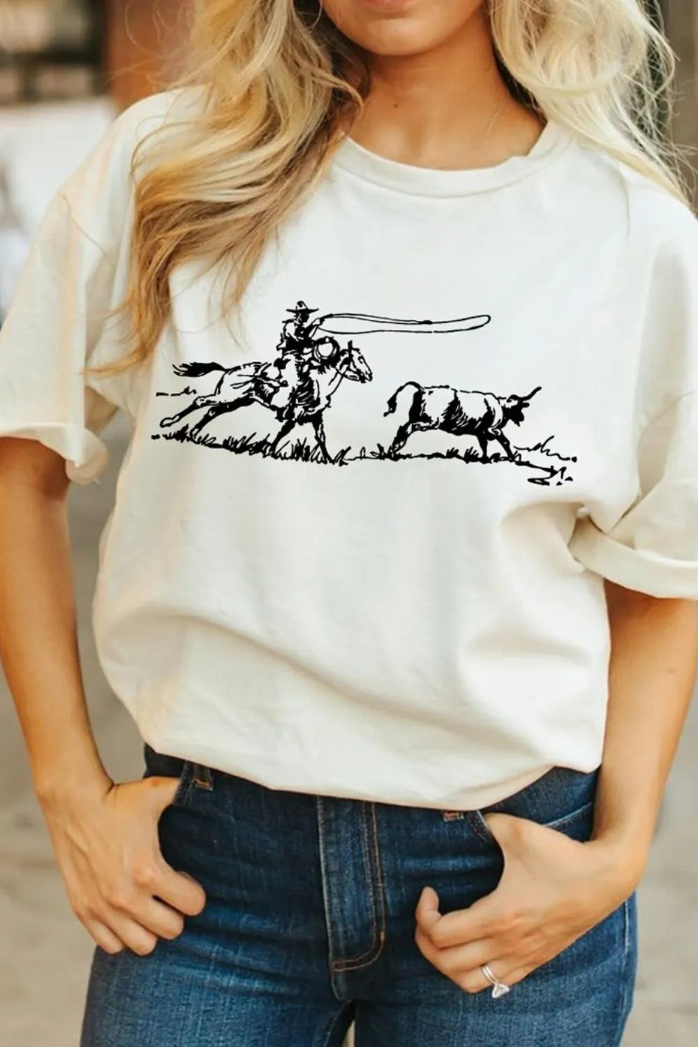 A person wearing blue jeans and a cowboy with horse chasing a calf graphic print t-shirt standing by a wood barn background