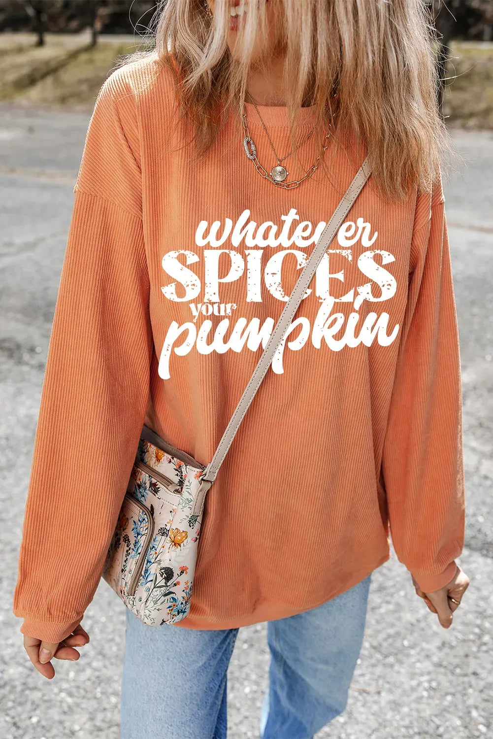 Person wearing an orange sweatshirt with text, holding a floral bag on a road.