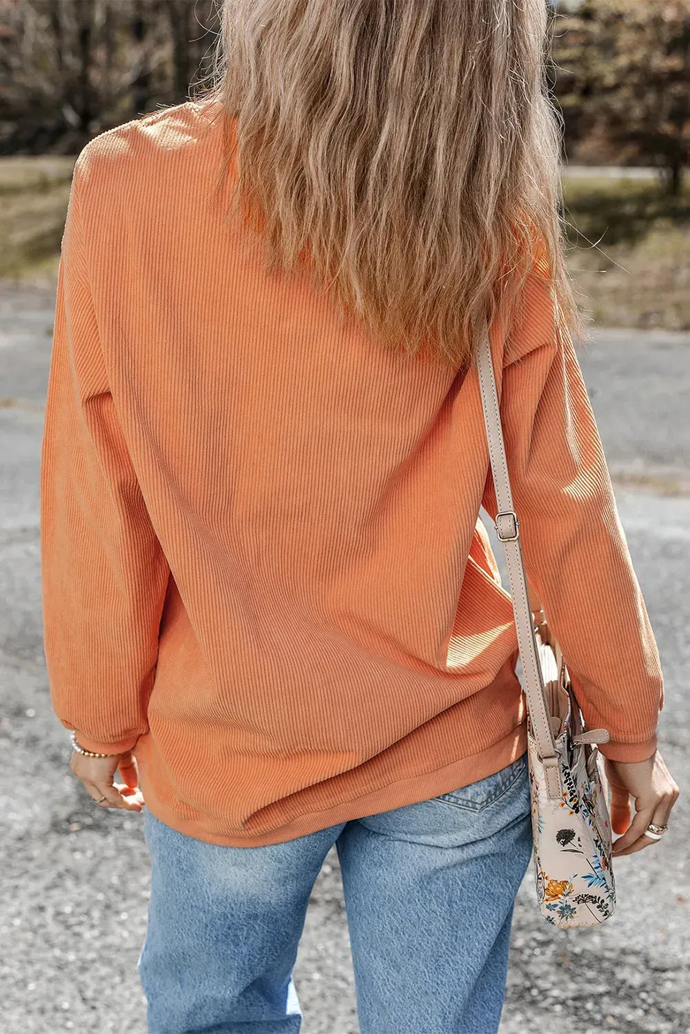 Person wearing an orange sweater and blue jeans, holding a floral bag outdoors.