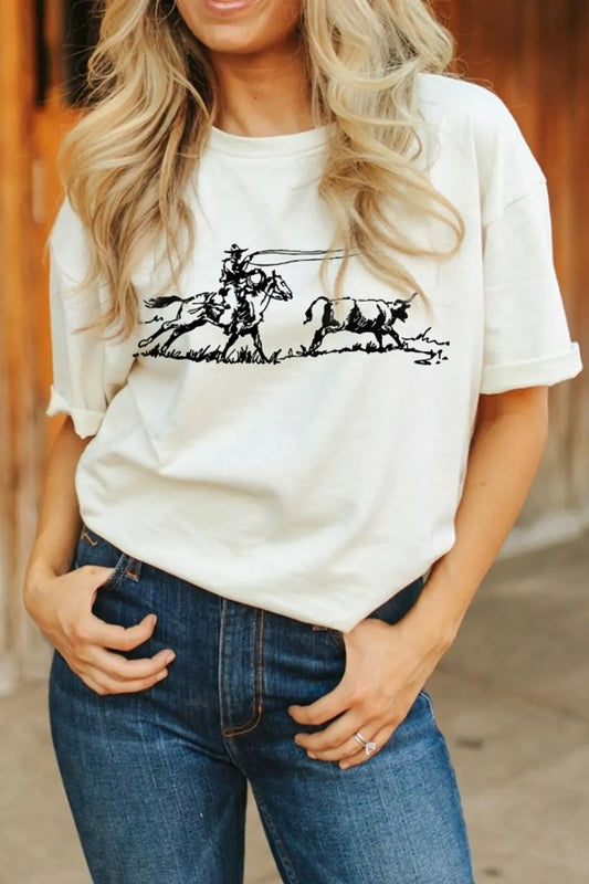 A person wearing blue jeans and a cowboy with horse chasing a calf graphic print t-shirt standing by a wood barn background
