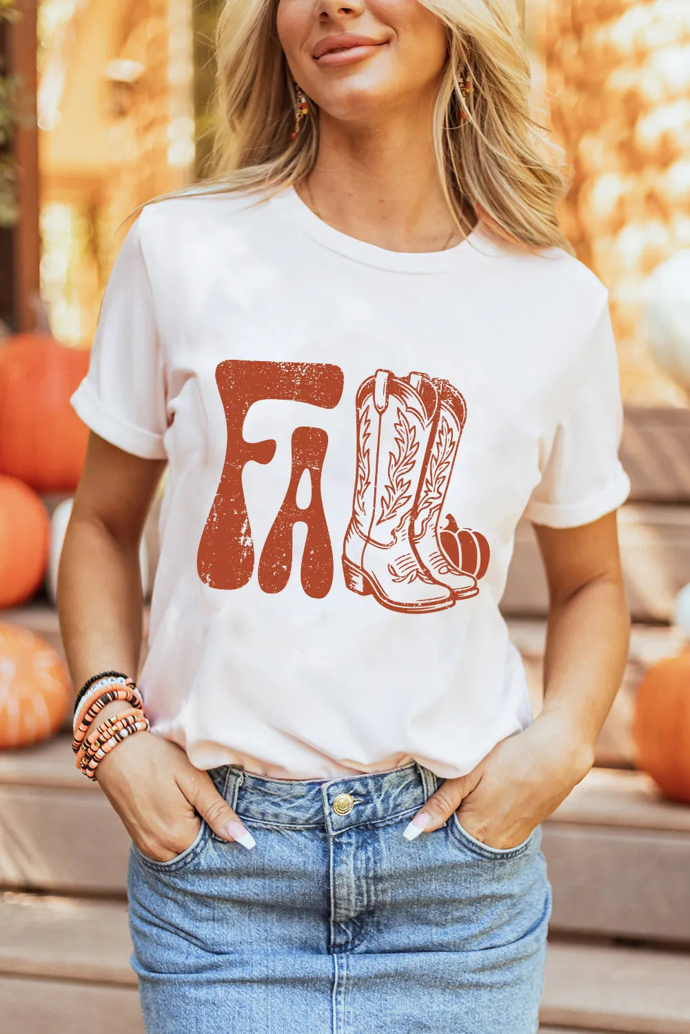 Woman wearing a white t-shirt with cowboy boots and 'FA' print, standing outdoors with pumpkins in the background.