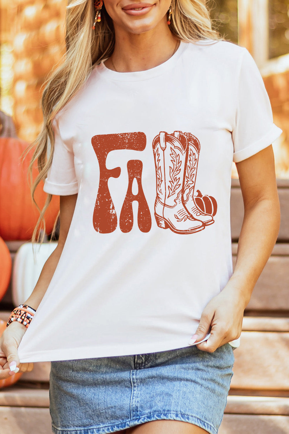 Women holding the front of a white tee shirt with the word Fall cowboy boots and pumpkin design and Jean mini skirt in front of a fall background.