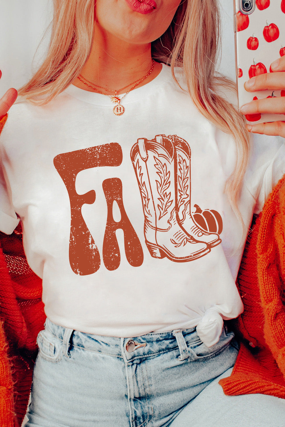 Women wearing a white cuffed Fall T-shirt with cowboy boots and pumpkin design and blue jeans holding a heart cell phone.