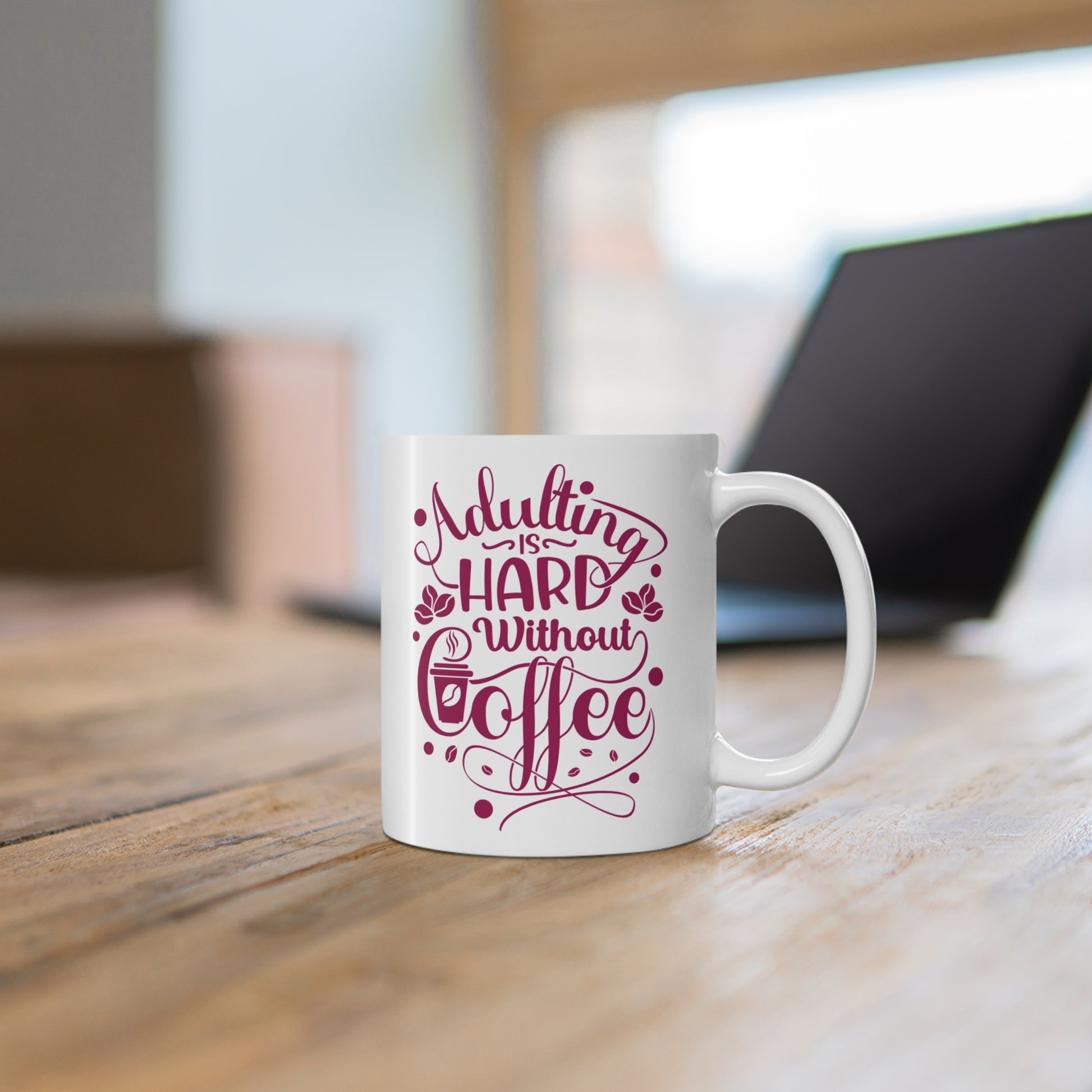 White mug with pink text 'Adulting is hard without coffee' on a wooden surface.