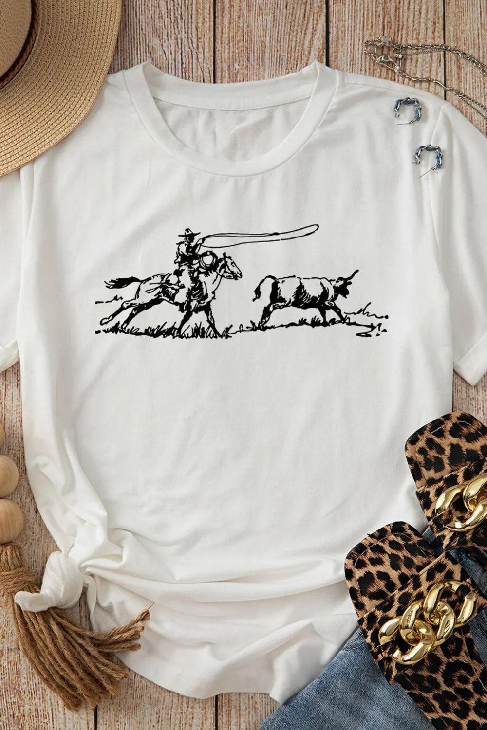T-shirt on wood table with horse chasing a calf graphic print t-shirt with a hat, leopard print sandals and folded blue jeans