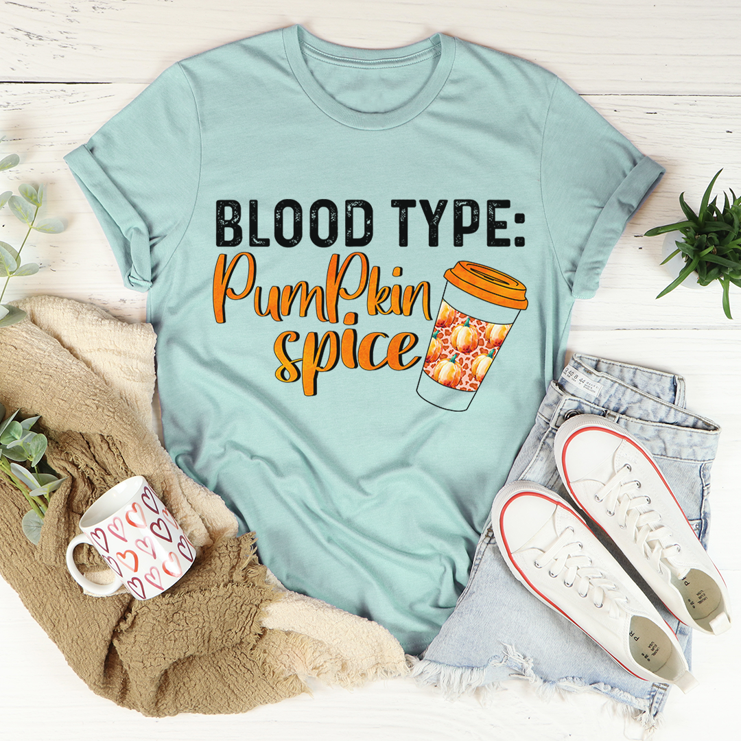 Light blue t-shirt with 'Blood Type: Pumpkin Spice' text and coffee cup graphic, surrounded by casual items on a wooden surface.