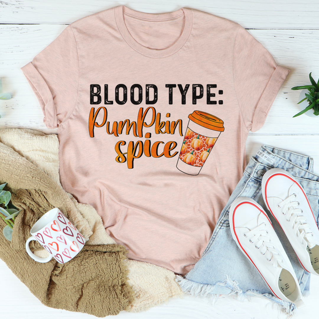 Pink t-shirt with 'Blood Type: Pumpkin Spice' text and coffee graphic, surrounded by casual items on a light wooden surface.