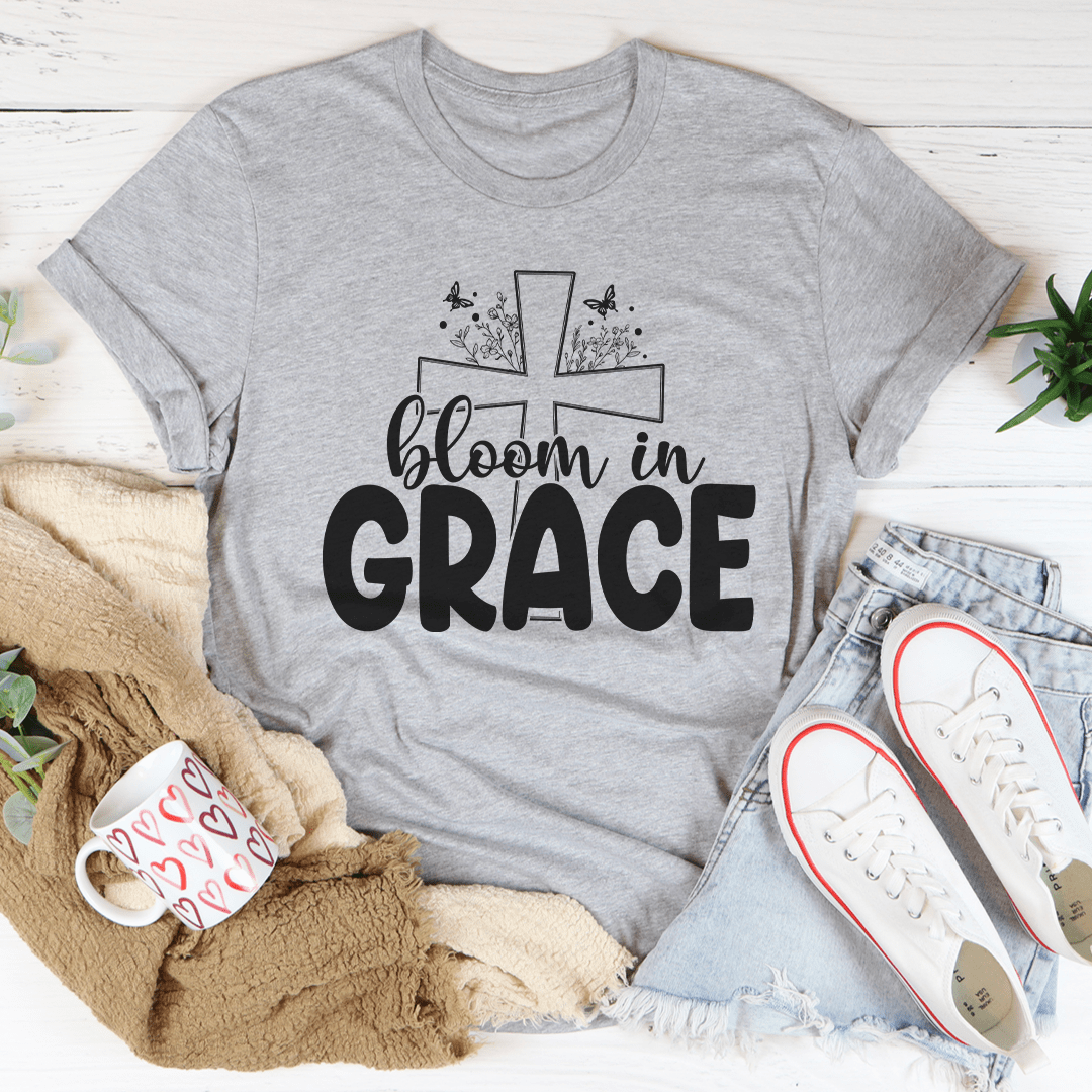 Gray t-shirt with 'bloom in grace' design on a white surface with casual items around
