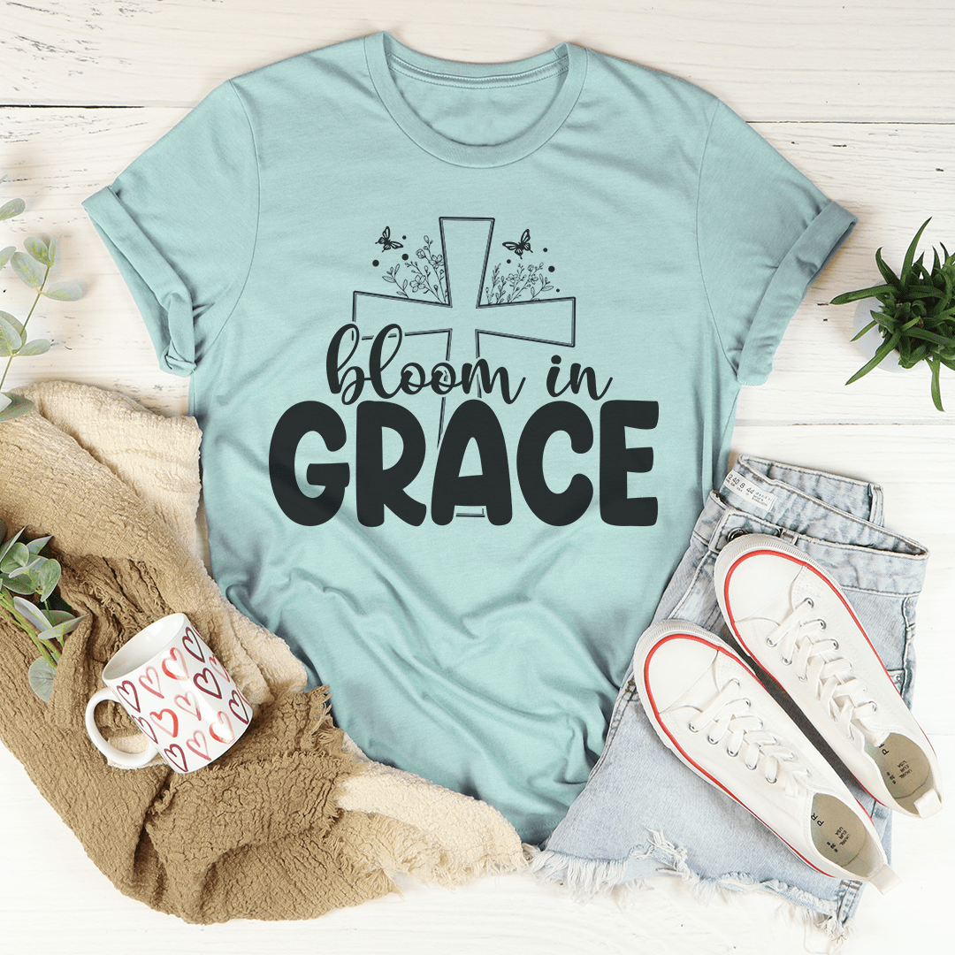 Light blue t-shirt with 'bloom in grace' text and cross design, surrounded by casual items on a light wooden surface.