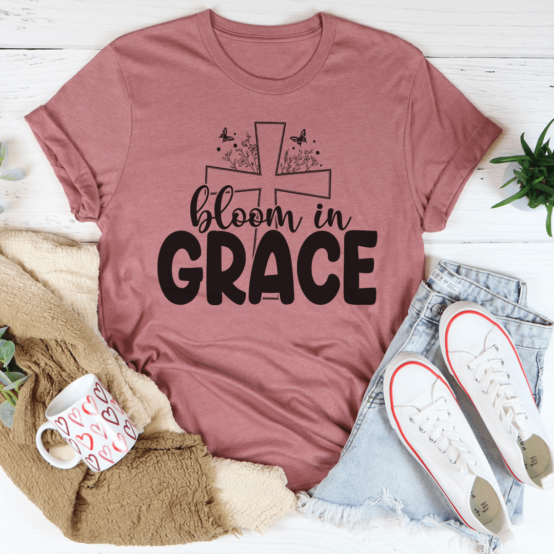 Mauve t-shirt with 'bloom in grace' text and cross design, surrounded by casual items on a light wooden surface.