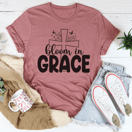Mauve t-shirt with 'bloom in grace' text and cross design, surrounded by casual items on a light wooden surface.