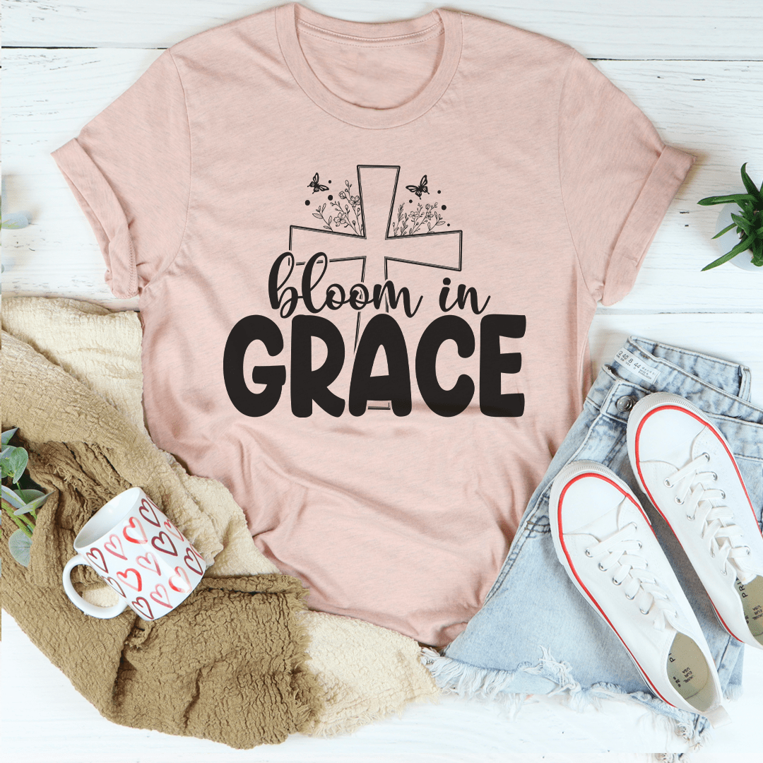 Pink t-shirt with 'bloom in grace' text on a white surface with casual items.