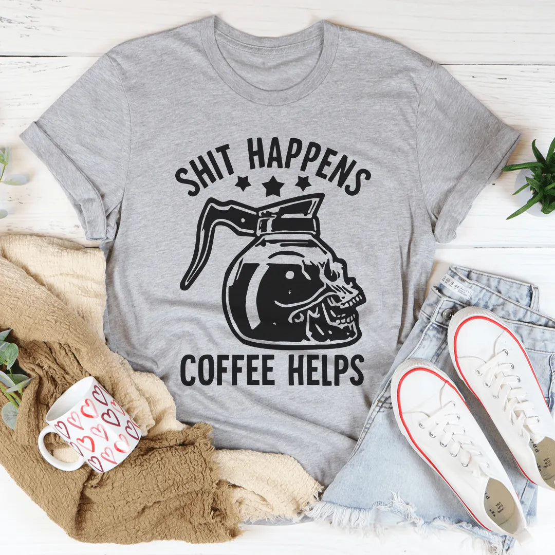 Gray t-shirt with 'Shit Happens Coffee Helps' graphic on a white surface with casual items.