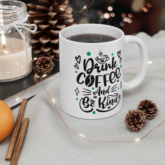 White mug with 'Drink Coffee And Be Kind' text on a table with decorative items.