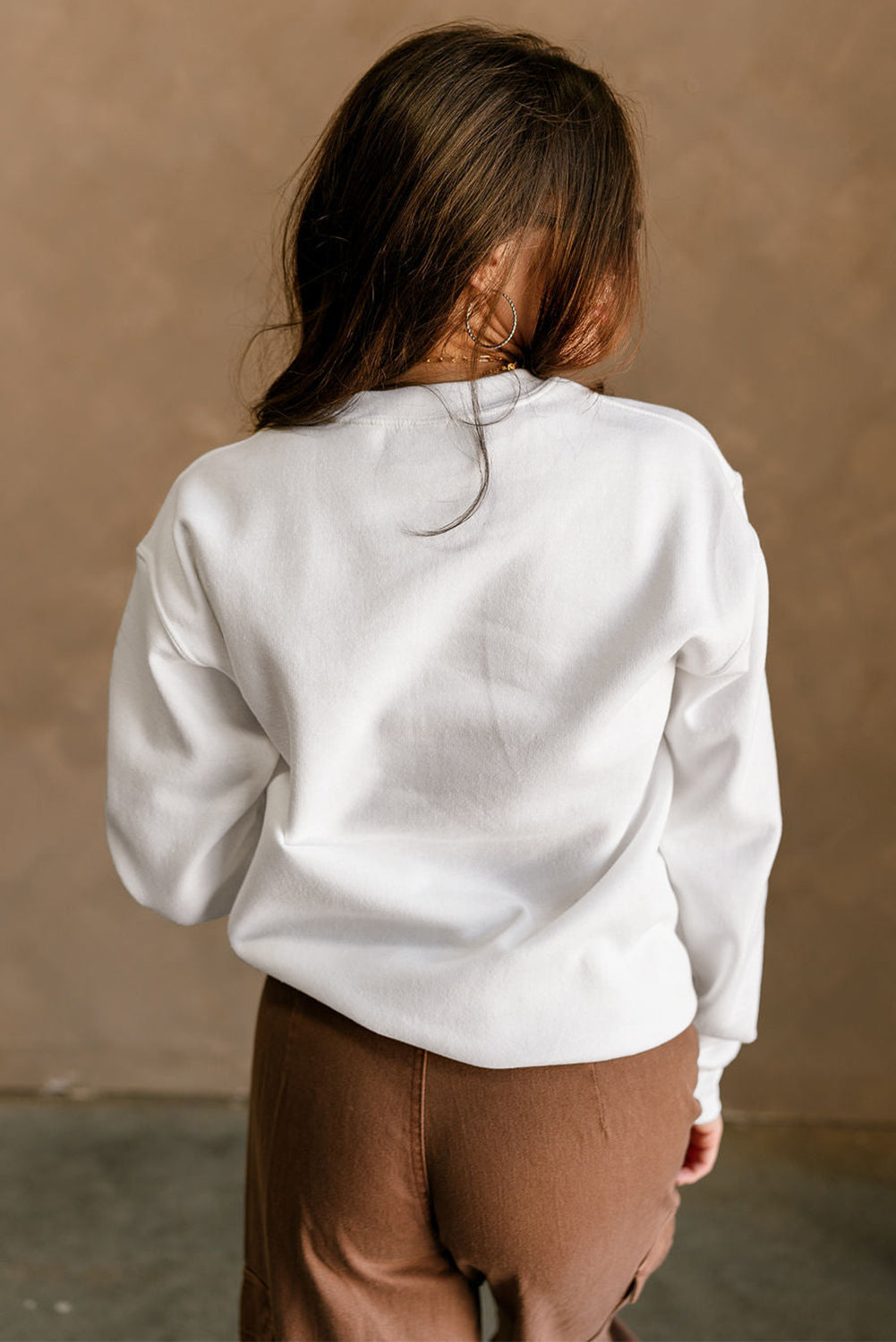 Person wearing a white sweatshirt and brown pants against a beige wall