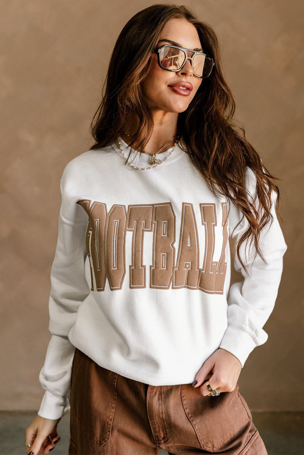 Woman wearing a white sweatshirt with 'FOOTBALL' text, sunglasses, and brown pants against a beige background