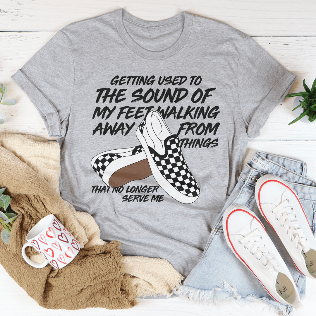 Gray t-shirt with checkered slip-on shoes graphic and text, placed on a surface with casual items like jeans and sneakers.