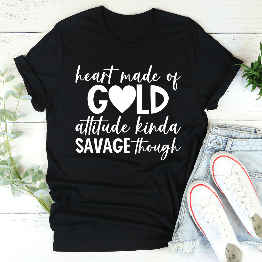 Black t-shirt with white text 'heart made of GOLD attitude kinda SAVAGE though' on a light wooden surface with shoes and plants.