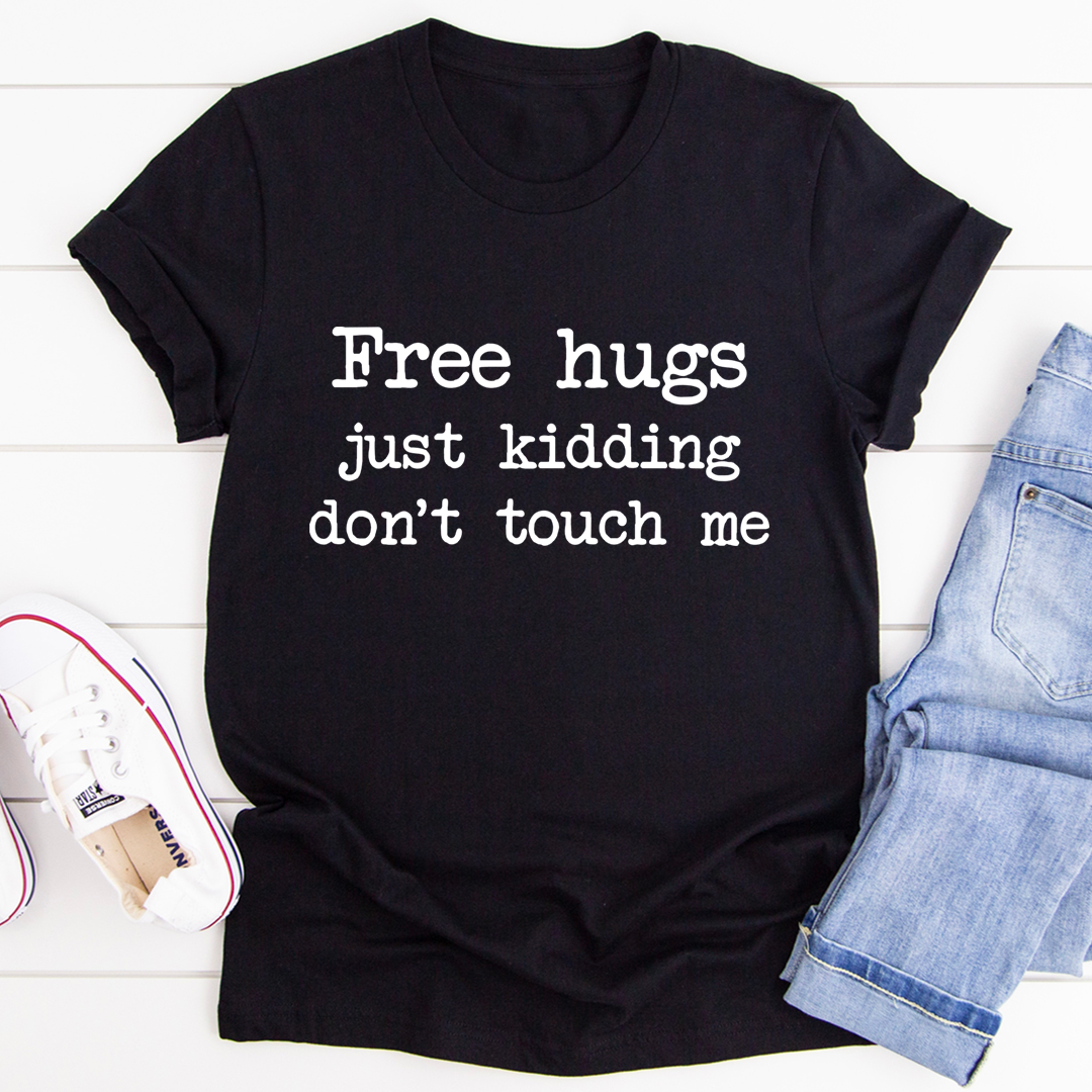 Black t-shirt with text 'Free hugs just kidding don't touch me' on a white wooden surface with jeans and a shoe.