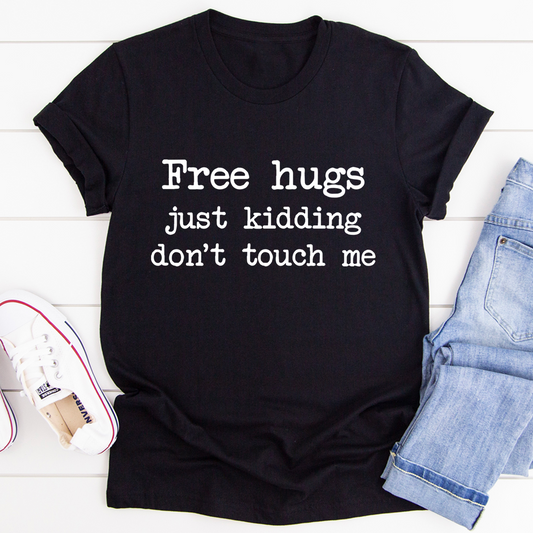 Black t-shirt with text 'Free hugs just kidding don't touch me' on a white wooden surface with jeans and a shoe.