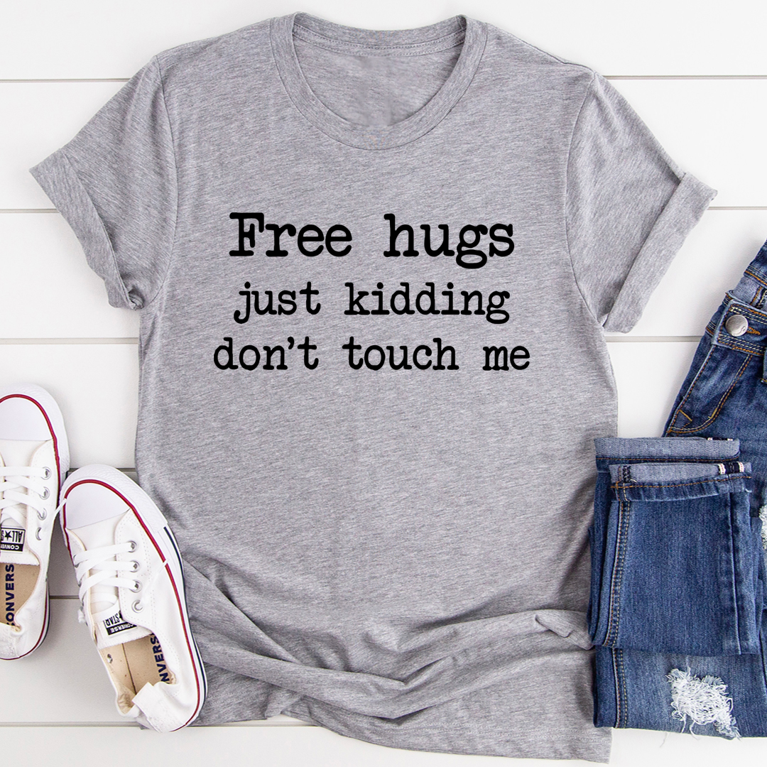 Gray t-shirt with 'Free hugs just kidding don't touch me' text, paired with jeans and sneakers on a light wooden surface.