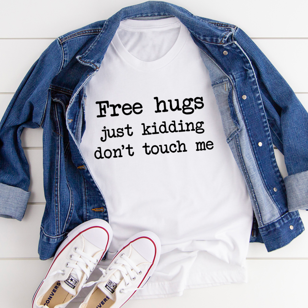 White t-shirt with text 'Free hugs just kidding don't touch me' on a white background with a denim jacket and white sneakers.