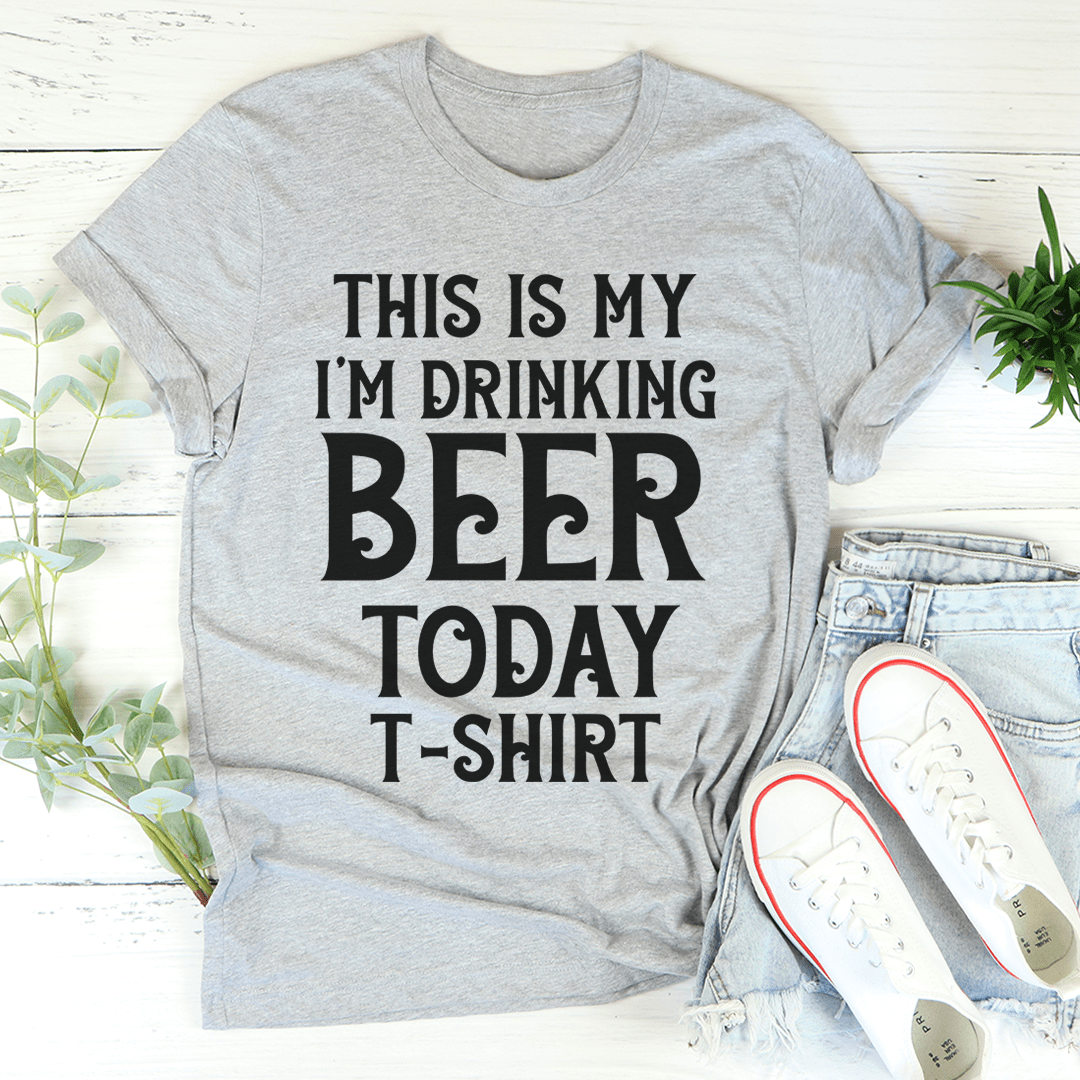 Gray t-shirt with 'This is my I'm drinking beer today' text, paired with jeans and shoes on a light wooden surface.
