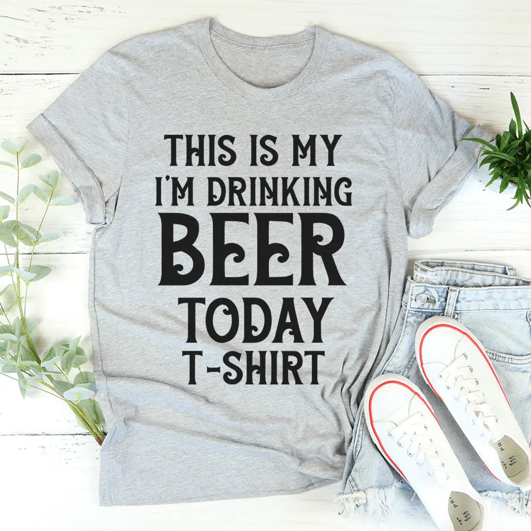 Gray t-shirt with 'This is my I'm drinking beer today t-shirt' text, paired with jeans and shoes on a light wooden surface.