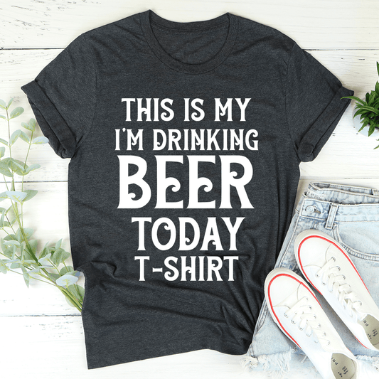 Dark gray t-shirt with white text 'This is my I'm drinking beer today t-shirt' on a light wooden surface.