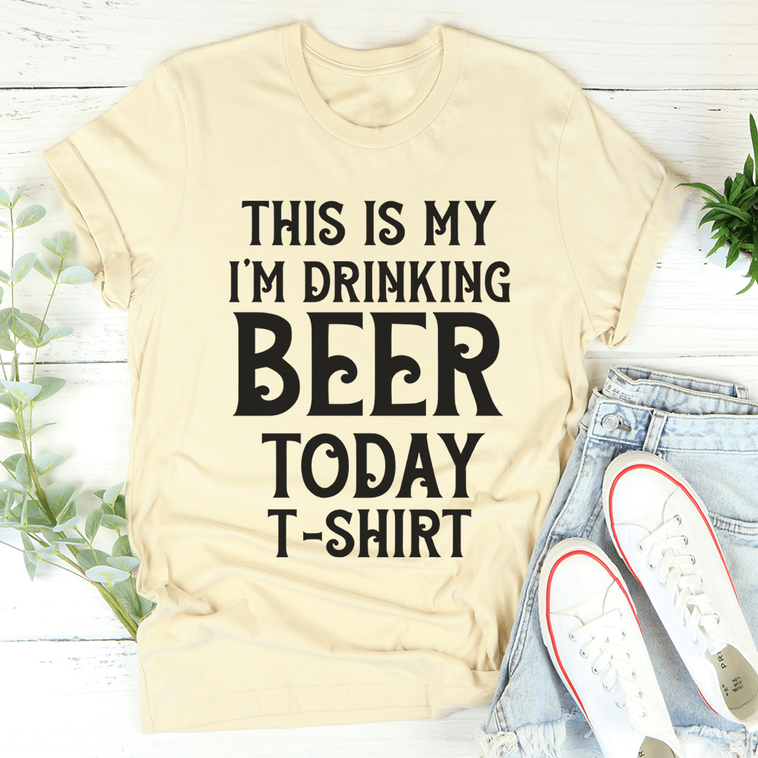 Beige t-shirt with text 'This is my I'm drinking beer today t-shirt' on a white wooden surface with jeans and shoes.