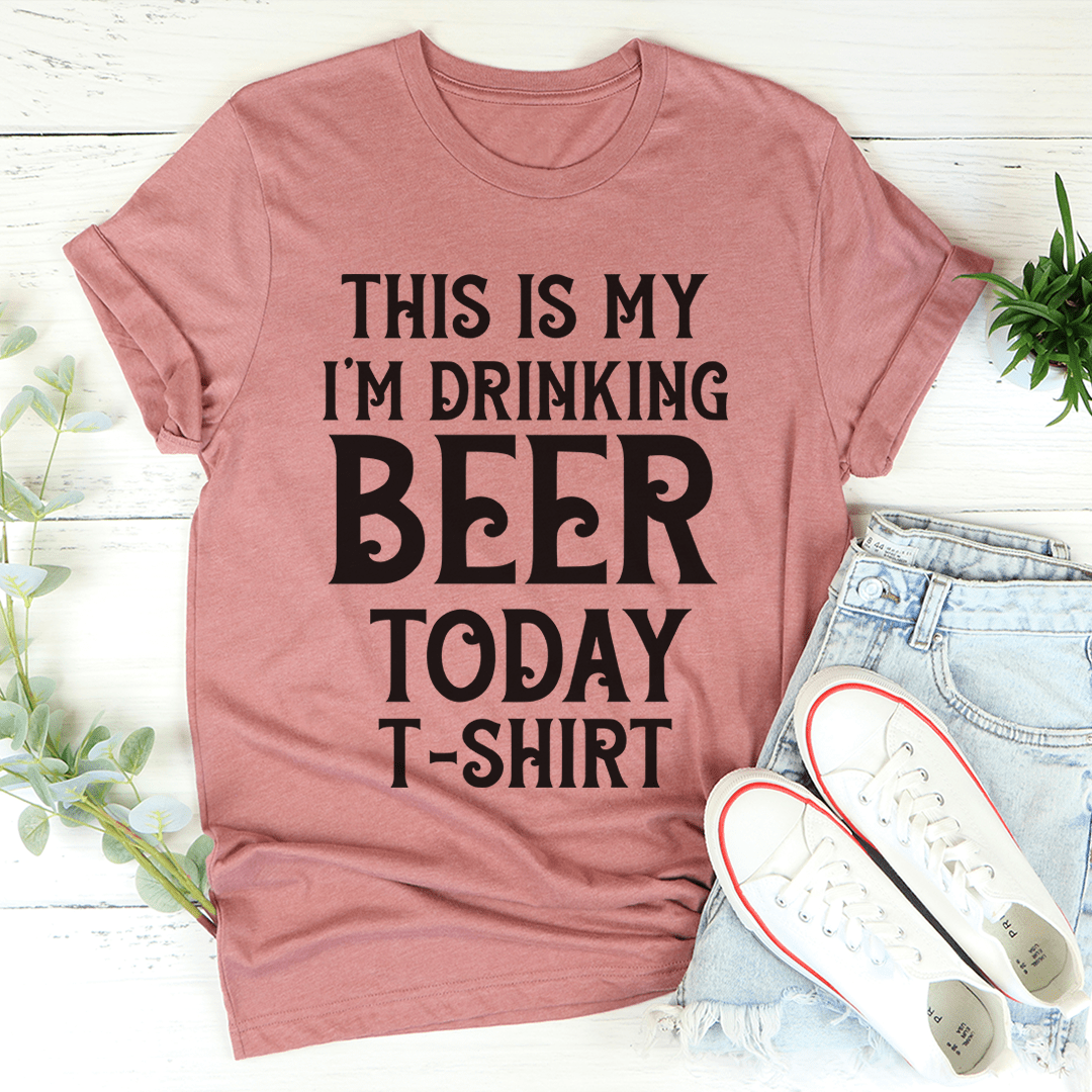 Pink t-shirt with 'This is my I'm drinking beer today' text, paired with jeans and sneakers on a light wooden surface.