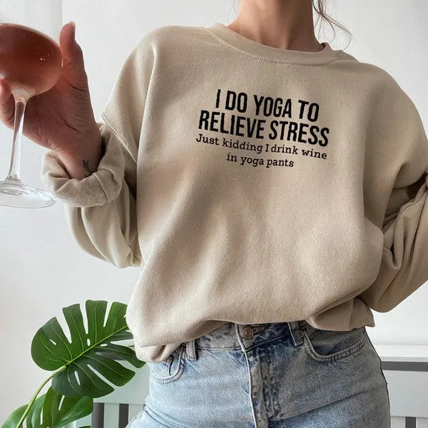 Person wearing a beige sweatshirt with text, holding a glass of wine, with a plant in the background.