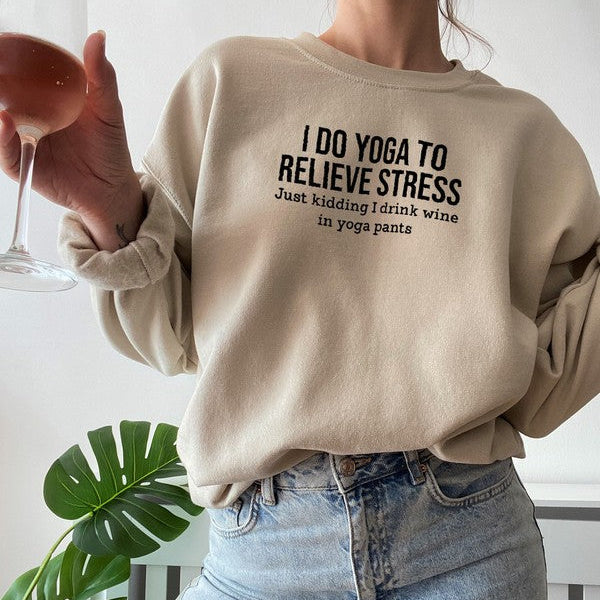 Person wearing a beige sweatshirt with text, holding a glass, standing in a room with a plant.