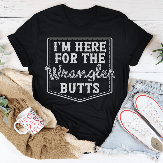 Black t-shirt with 'I'm here for the Wrangler Butts' text on a wooden surface with casual items.