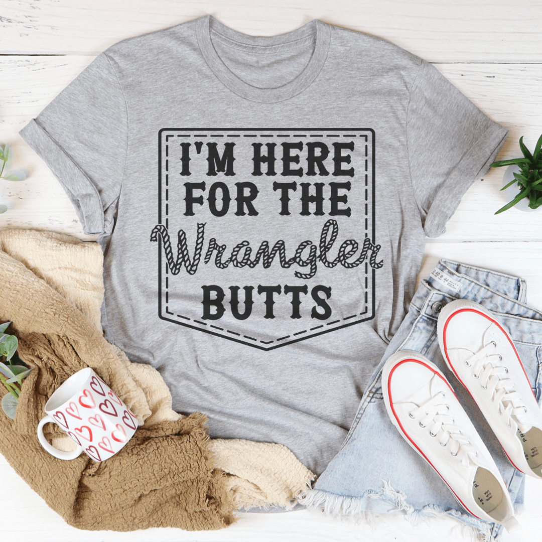 Gray t-shirt with 'I'm here for the Wrangler Butts' text, surrounded by casual items on a light wooden surface.