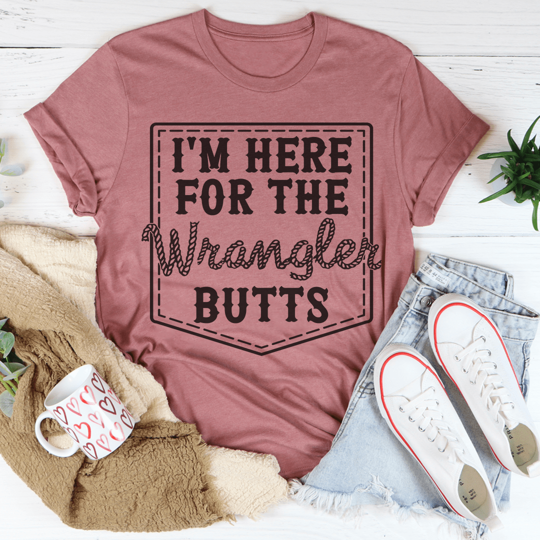 Mauve t-shirt with 'I'm here for the Wrangler Butts' text, placed on a white surface with casual items around.