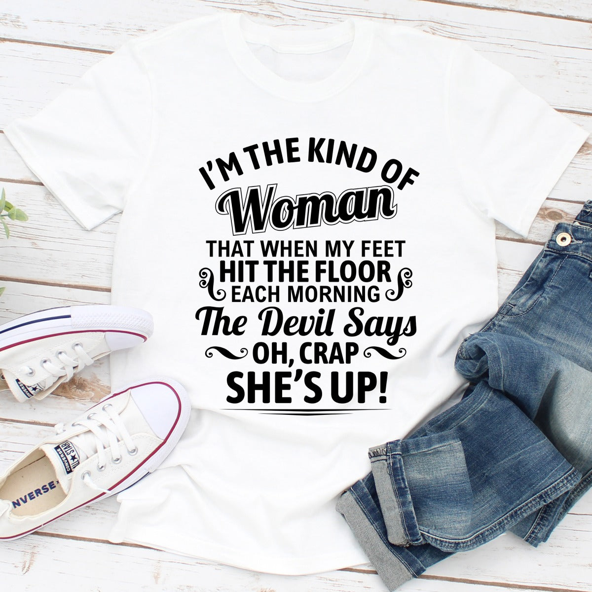 White t-shirt with humorous text design, paired with jeans and sneakers on a wooden surface.