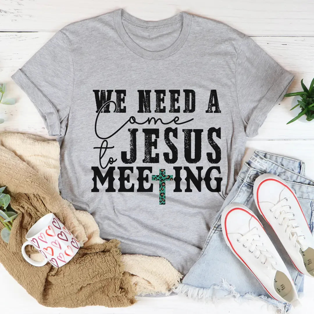 Gray t-shirt with 'We Need a Jesus Meeting' text on a white surface with casual items.