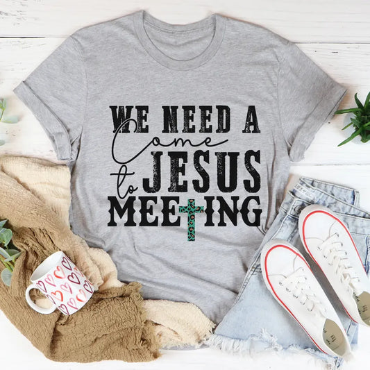 Gray t-shirt with 'We Need a Jesus Meeting' text on a white surface with casual items.