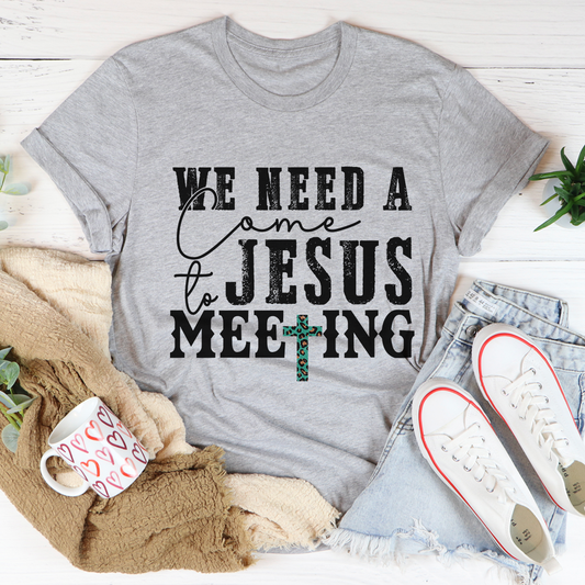 Gray t-shirt with 'We need a Jesus meeting' text on a white surface with casual items around.