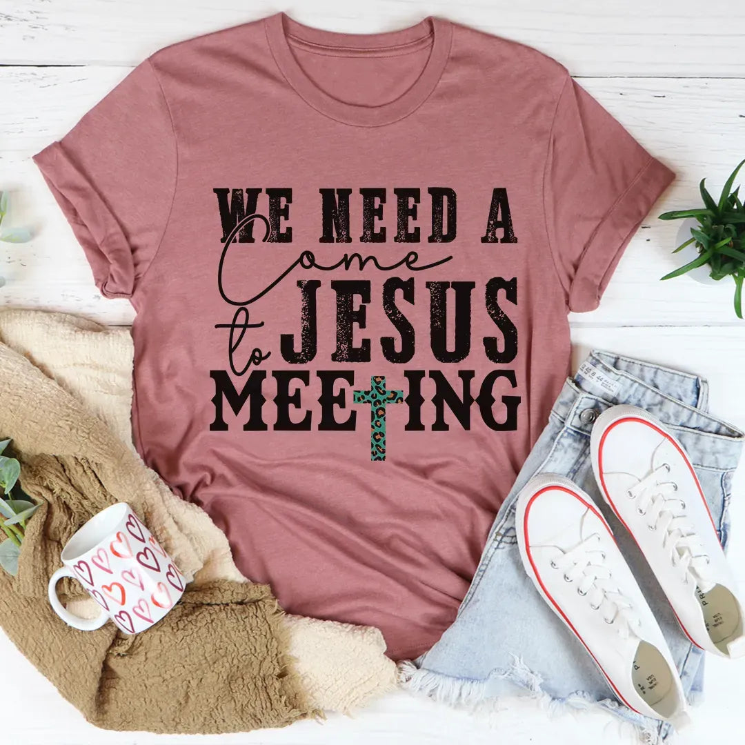 Mauve t-shirt with 'We need a come to Jesus meeting' text and cross graphic, surrounded by casual items on a light wooden surface.