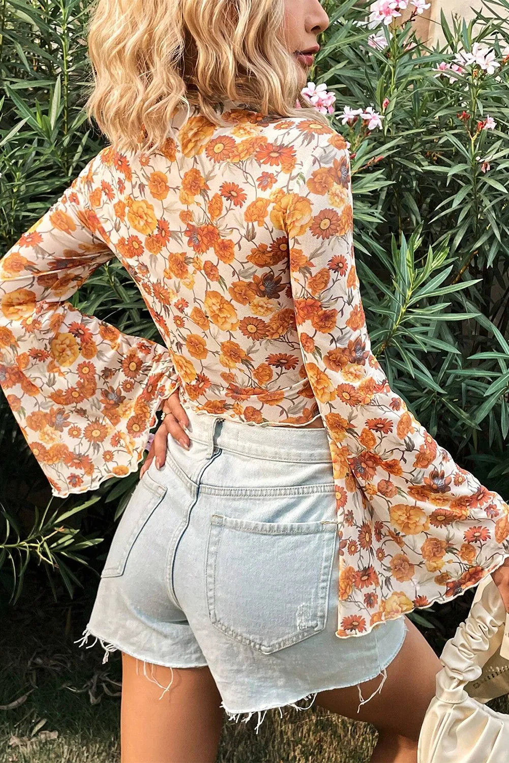 Person wearing a floral blouse with long sleeves and denim shorts standing in front of green foliage.