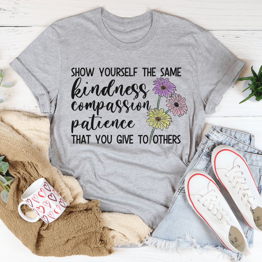 Gray t-shirt with motivational quote and flowers, surrounded by casual items on a light wooden surface.