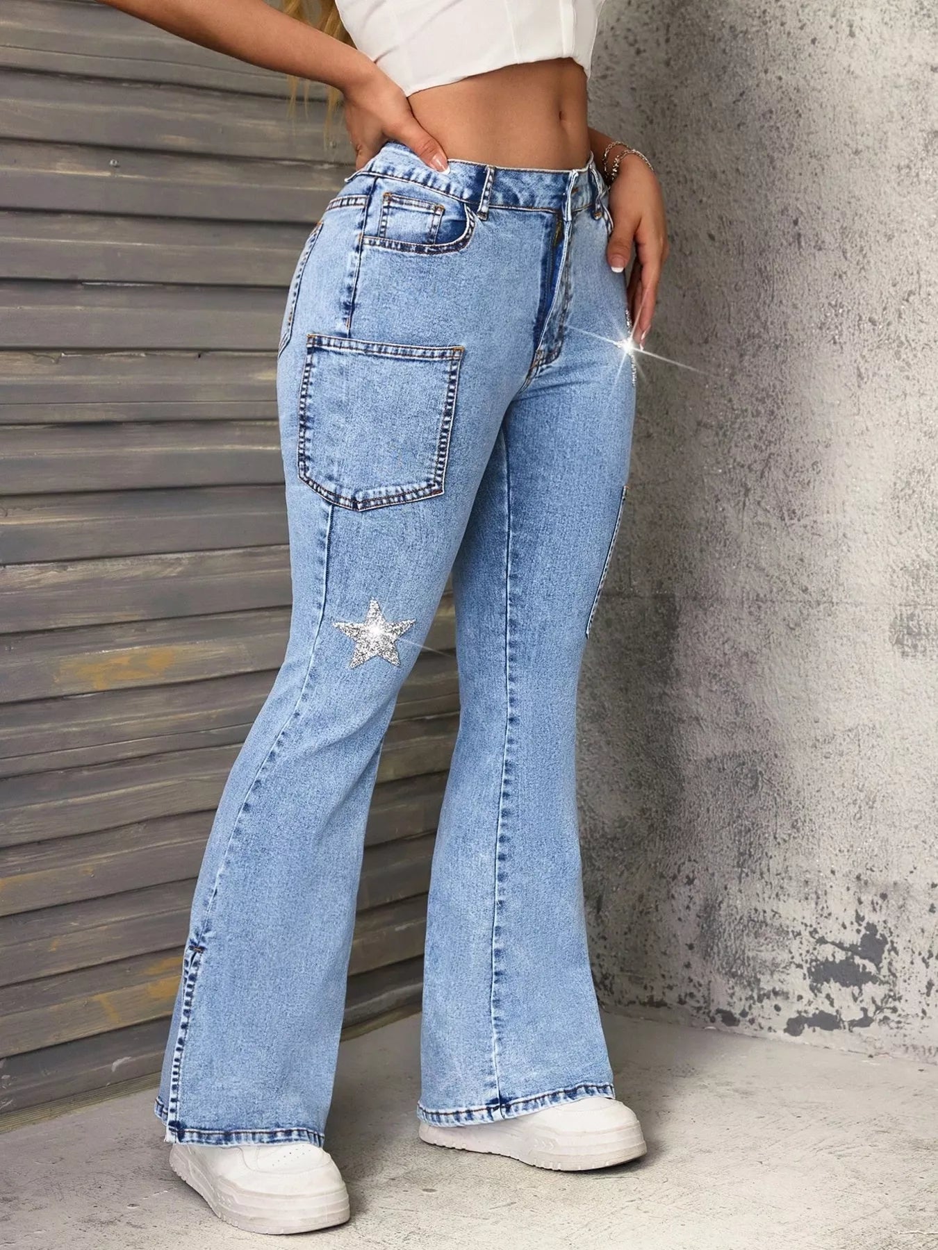 Lady wearing slit star flare leg blue denim jeans paired with a white tank top and white tennis shoes, with walls visible in the background.
